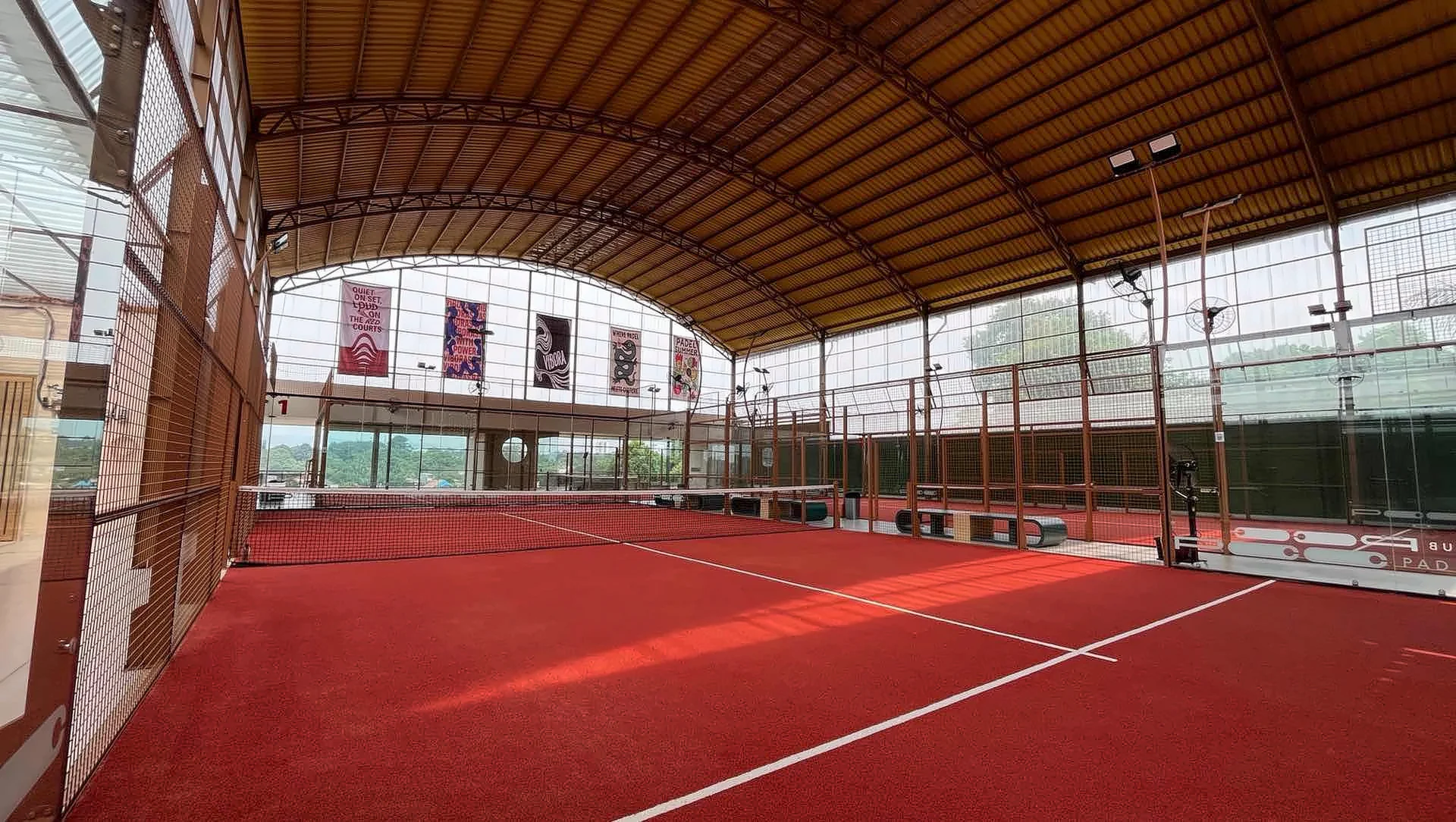 PCC Bogor: terracotta padel courts under a high arched timber roof, glass walls all round